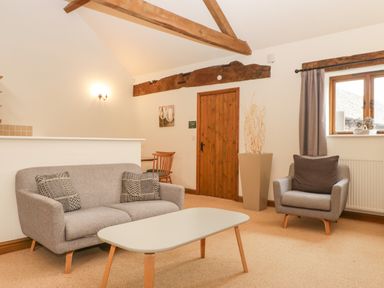 A living room with a sofa, armchair, coffee table, wooden door, window with curtain, and exposed wooden beams at Honeysuckle Cottage in Stanford Bishop near Bromyard