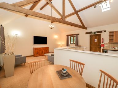 An open living and dining area with wooden beams a round wooden table grey armchairs a wall mounted television and a kitchen area at Honeysuckle Cottage in Stanford Bishop near Bromyard