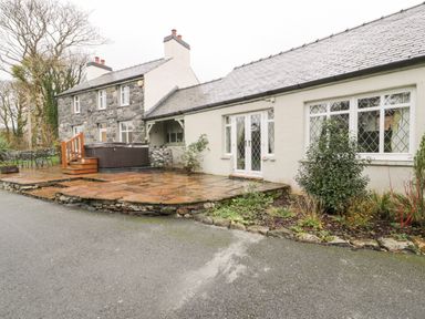 Exterior view of a stone and white painted house with a paved patio area hot tub and outdoor seating at Lakeside Cottage in Llanrug