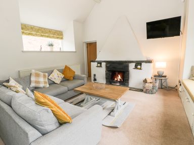 A living room with two grey sofas cushions a wooden coffee table a fireplace with a fire a wall-mounted television a window with a yellow curtain and a side table with a lamp at Lakeside Cottage in Llanrug