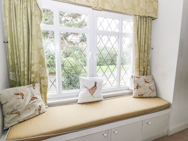 A window seat with three bird-themed cushions under a diamond-pane window with green floral curtains at Lakeside Cottage in Llanrug