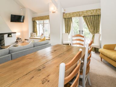 A dining area with a wooden table and chairs next to a living room with grey and yellow sofas and windows with green curtains at Lakeside Cottage in Llanrug