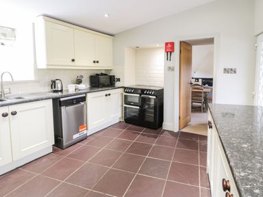 A kitchen with white cabinets a dishwasher a kettle a microwave a stove and a doorway leading to a dining area at Lakeside Cottage in Llanrug