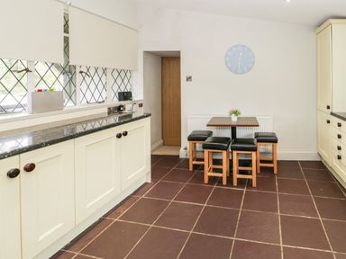 A kitchen with cream cabinets a small dining table and stools near a wall clock at Lakeside Cottage in Llanrug