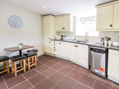 A kitchen with a small dining table and stools a wall clock and white cabinets with a dishwasher at Lakeside Cottage in Llanrug