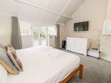 A bedroom with a bed and pillows towels on the bed a white dresser a TV mounted on the wall and glass doors at Lakeside Cottage in Llanrug