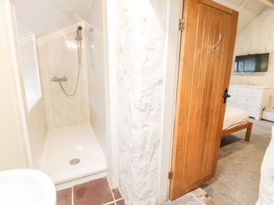 A bathroom with a shower and sink next to a wooden door leading to a bedroom with a bed dresser and wall mounted tv at Lakeside Cottage in Llanrug