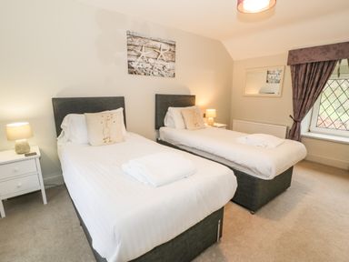 A bedroom with two single beds side by side each with white bedding and a pillow with a bird design two bedside tables with lamps a wall mirror and window with purple curtains at Lakeside Cottage in Llanrug