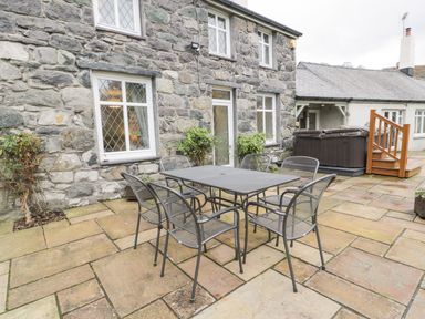 An outdoor stone patio with metal dining table and chairs next to a stone building with windows at Lakeside Cottage in Llanrug