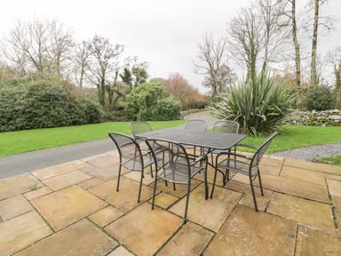 An outdoor patio with a metal table and six chairs surrounded by grass and trees at Lakeside Cottage in Llanrug