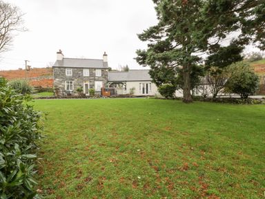 A stone house with two chimneys and an attached white building surrounded by a green lawn and trees at Lakeside Cottage in Llanrug