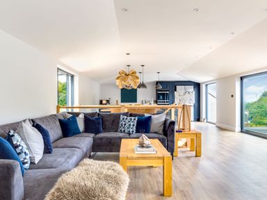 A living room with a grey sectional sofa and wooden tables open to a kitchen with dark cabinetry at Breakers View in Polzeath