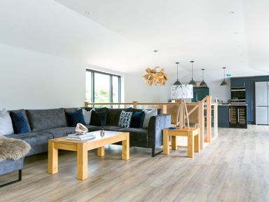 A living room with a gray sectional sofa and wooden coffee table next to a kitchen with blue cabinets at Breakers View in Polzeath
