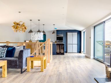 An open living space with a gray sofa and wooden tables next to a kitchen with blue cabinets and stainless steel refrigerator at Breakers View in Polzeath