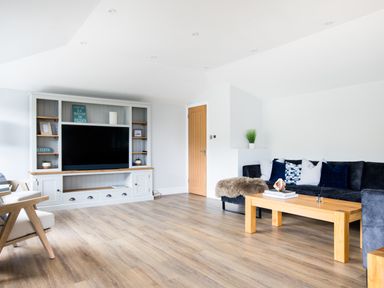 A living room with wooden flooring a large black sofa wooden coffee table a white entertainment unit with a television and wooden door at Breakers View in Polzeath