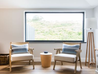 Two wooden armchairs with cushions and a small round table between them in front of a large window in a living room at Breakers View in Polzeath