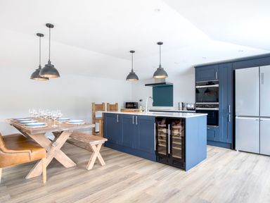 A kitchen with blue cabinets an island with wine coolers wooden dining table set with glassware pendant lights and stainless steel appliances at Breakers View in Polzeath