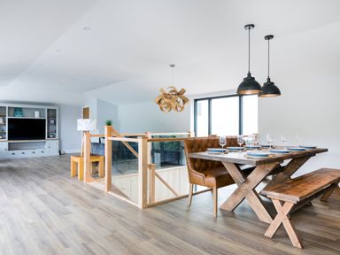 A dining area with a wooden table set with plates and glasses brown leather bench seating and wooden bench seating next to a staircase with glass and wood railing in an open living space at Breakers View in Polzeath