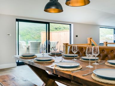 A dining table set with plates and wine glasses in a room with large windows and a leather bench at Breakers View in Polzeath