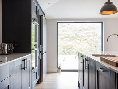 A kitchen with dark cabinets a refrigerator oven toaster and a large window showing an outdoor view at Breakers View in Polzeath
