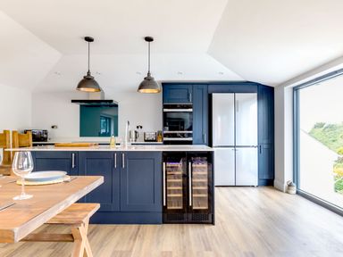 A kitchen with blue cabinets a wooden dining table and large window at Breakers View Polzeath