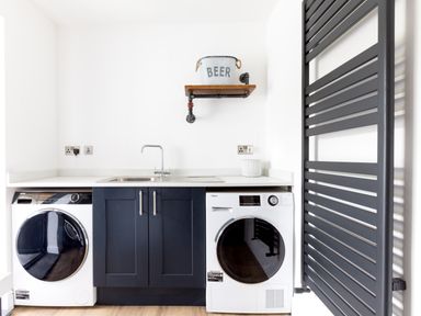 A laundry room with a washing machine and dryer under a countertop with a sink and faucet and a wall shelf holding a bucket labeled beer at Breakers View in Polzeath