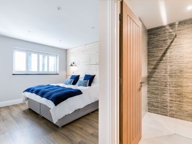A bedroom with a bed with blue bedding and pillows next to a bathroom with a wooden door and a tiled shower at Breakers View in Polzeath