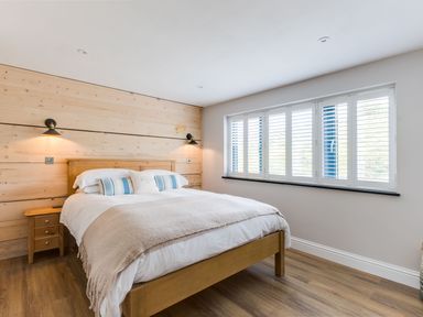 A bedroom with a wooden bed frame wooden panel wall bedside tables and a window with white shutters at Breakers View in Polzeath