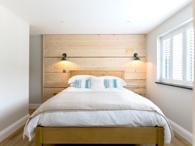 A bed with pillows and a blanket between two wall lamps and a window with shutters at Breakers View Polzeath