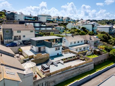 A neighborhood with multiple houses on a hillside including a modern two-story house with a patio and outdoor furniture at Breakers View in Polzeath
