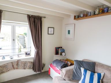 A corner room with a window with curtains a sofa with cushions a small shelf with books and framed pictures at 1 The Old Brewhouse in Dartmouth