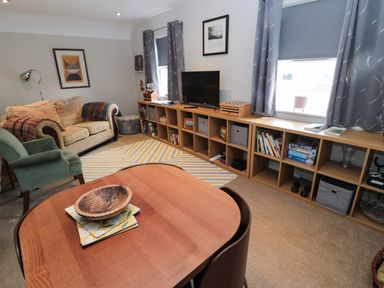 A living room with a wooden table in the foreground a sofa and armchair against the wall a rug on the floor and shelving units with a TV at 60B Castlegate in Berwick-Upon-Tweed