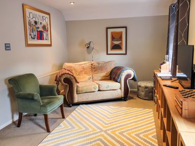 A sitting room with a patterned sofa, a green armchair, a wooden shelf unit, framed artwork on the walls, and a chevron-patterned rug at 60B Castlegate in Berwick-Upon-Tweed