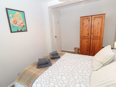 A bedroom with a bed with white patterned bedding and cushions wooden wardrobe and a framed painting of flowers on the wall at 60B Castlegate in Berwick-Upon-Tweed