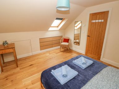 A bedroom with a bed two folded towels a wooden door a wicker chair a wall mirror skylight window and a wooden table at Green Grove Barn in Peniel near Carmarthen