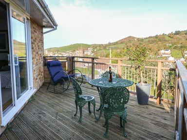 A balcony with a round metal table two chairs a rocking chair and a potted plant overlooking hills and houses at Hillsview in Combe Martin