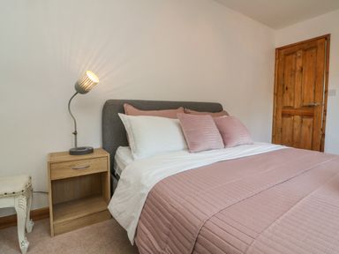 A bedroom with a gray headboard bed with pink and white bedding and wooden nightstand with a lamp near a wooden door at Hillsview in Combe Martin