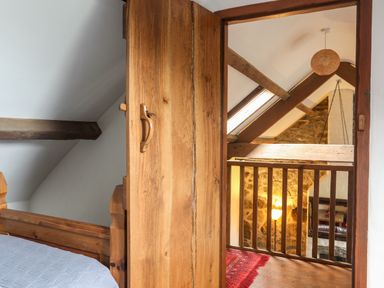 A wooden door opening to a loft area with wooden beams and a railing overlooking a stone wall and lamp at Tir Eironwy in Dinas near Nefyn