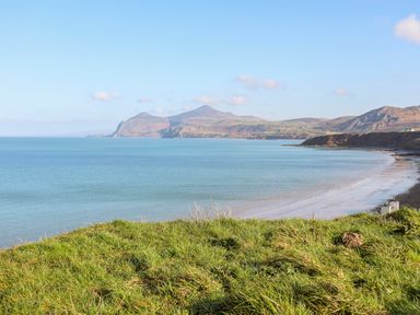 A coastal landscape with green grass in the foreground and a sandy beach along a blue sea with hills in the background at Tir Eironwy in Dinas near Nefyn