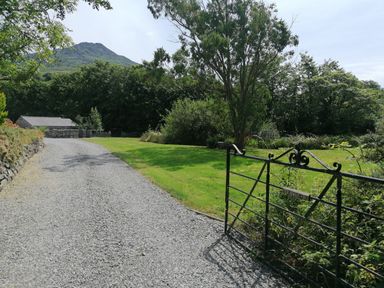 A gravel driveway with a metal gate and a stone wall beside trees and a mountain in the background at Tir Eironwy in Dinas near Nefyn