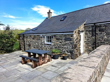 An outdoor patio area with a wooden picnic table and benches beside a stone house with a tiled roof and potted plants at Tir Eironwy in Dinas near Nefyn