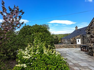 A stone house with a slate roof next to a paved patio and flowering bushes at Tir Eironwy Dinas near Nefyn