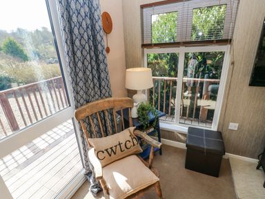 A corner of a room with a wooden chair with cushions a blue side table with a lamp and plant next to windows and a small black ottoman at 1 Hornbeam Lodge in Narberth