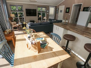 A dining area with a wooden table and blue chairs next to a kitchen island with bar stools and a living room with a sofa and wall-mounted TV at 1 Hornbeam Lodge in Narberth