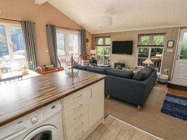 An open living room and kitchen area with a washing machine wooden table gray sofa and a wall-mounted television at 1 Hornbeam Lodge in Narberth