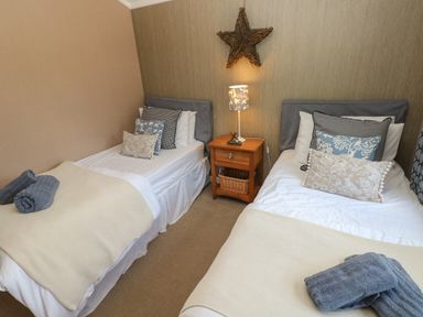 A bedroom with two single beds with throw pillows and folded towels on blankets with a wooden nightstand and a lamp in between at 1 Hornbeam Lodge in Narberth