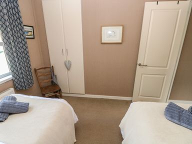 A bedroom with two single beds, a wooden chair, a white wardrobe, a door, a window with patterned curtains, and a framed picture on the wall at 1 Hornbeam Lodge Narberth