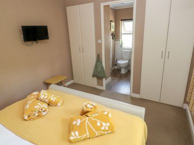 A bedroom with a bed covered in yellow bedding and towels two white wardrobes a small wall-mounted TV and an ensuite bathroom at 1 Hornbeam Lodge in Narberth