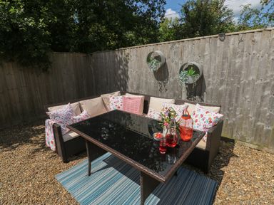 An outdoor seating area with a wicker corner sofa with cushions and a glass-top table on a striped rug at 1 Hornbeam Lodge in Narberth