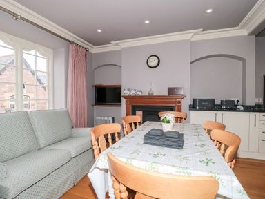 A dining room with a table set with placemats and a flower pot surrounded by wooden chairs a patterned sofa a fireplace with decorative jars and a clock above it and kitchen appliances on a counter at The Pound in Porlock